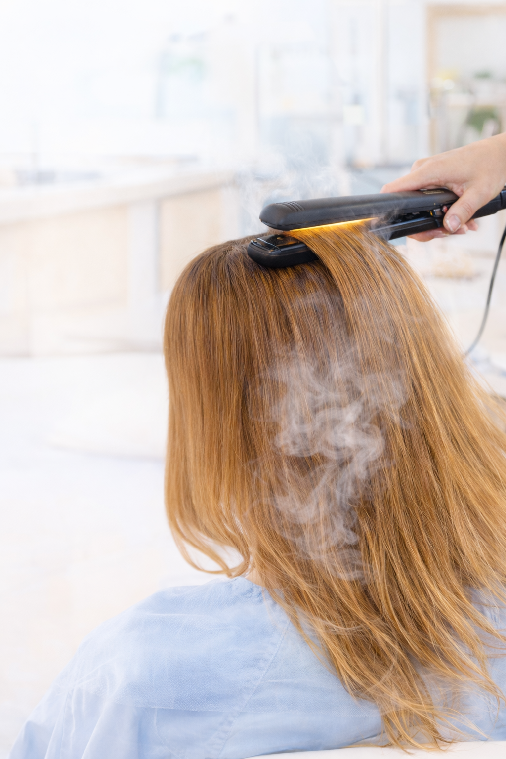Close-up of a flat iron smoothing brown hair in a salon, with steam visible during a keratin treatment heat activation process.